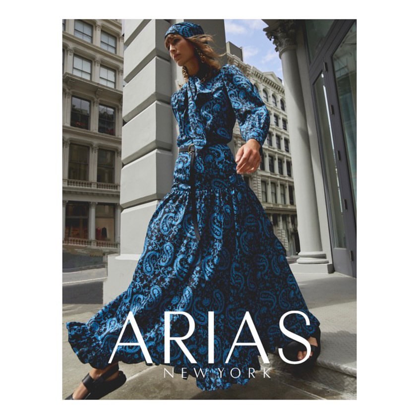 A fashionable adult female strides across an urban street wearing a matching blue paisley blouse, skirt, and head covering.
