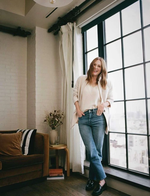 Stylish woman with red hair posing in an apartment interior with a couch, vase with flowers, books, and a window with a city view.