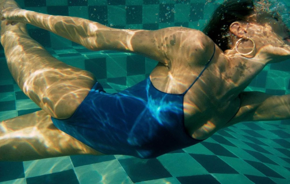 Underwater shot of a woman in a blue swimsuit swimming in a pool with green and white checkered tiles and bubbles rising in the water.