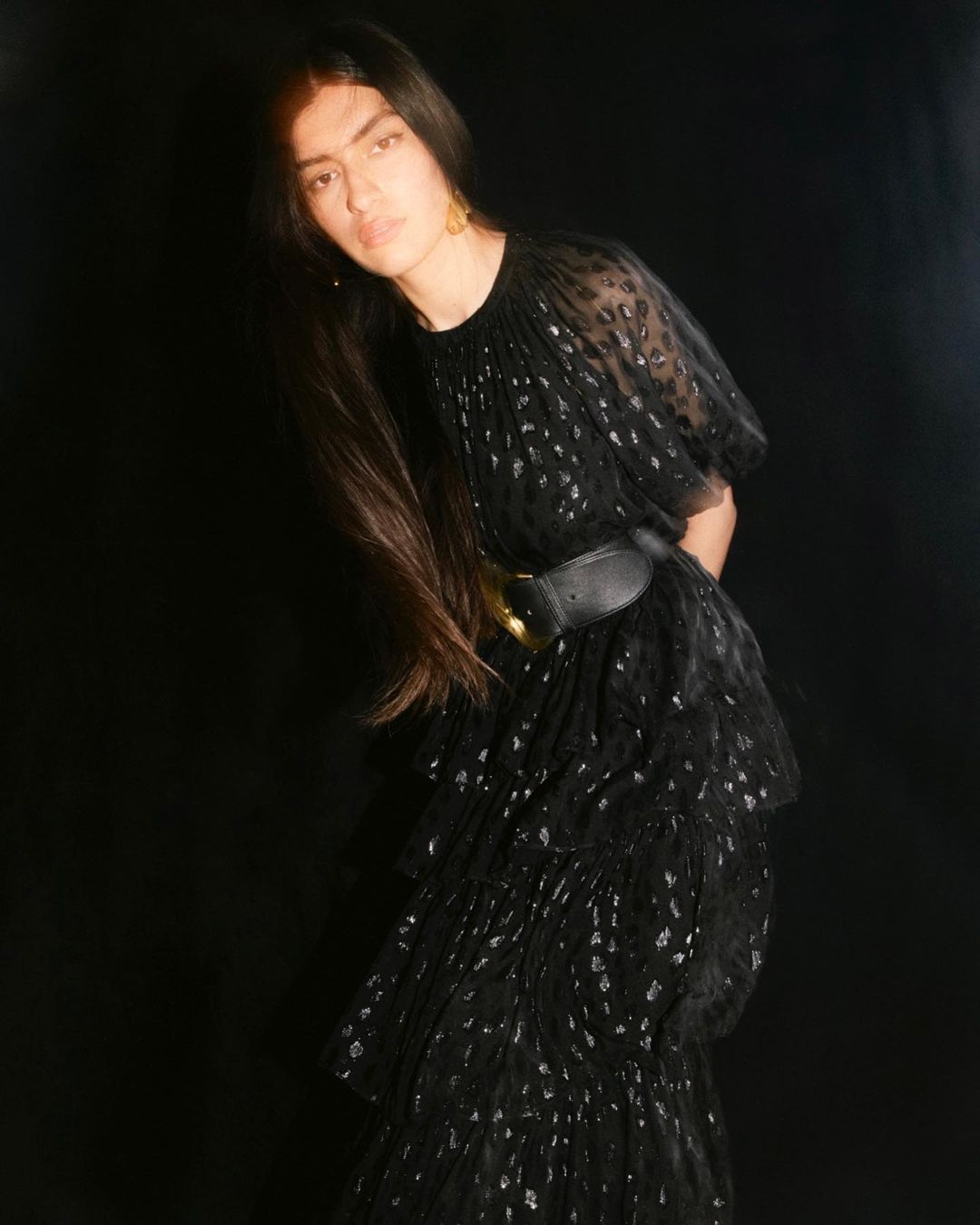 A woman with long dark hair poses in a black, tiered, semi-sheer dress cinched with a wide black belt against a dark background.