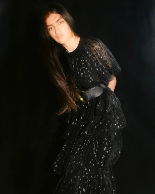 A woman with long dark hair poses in a black, tiered, semi-sheer dress cinched with a wide black belt against a dark background.