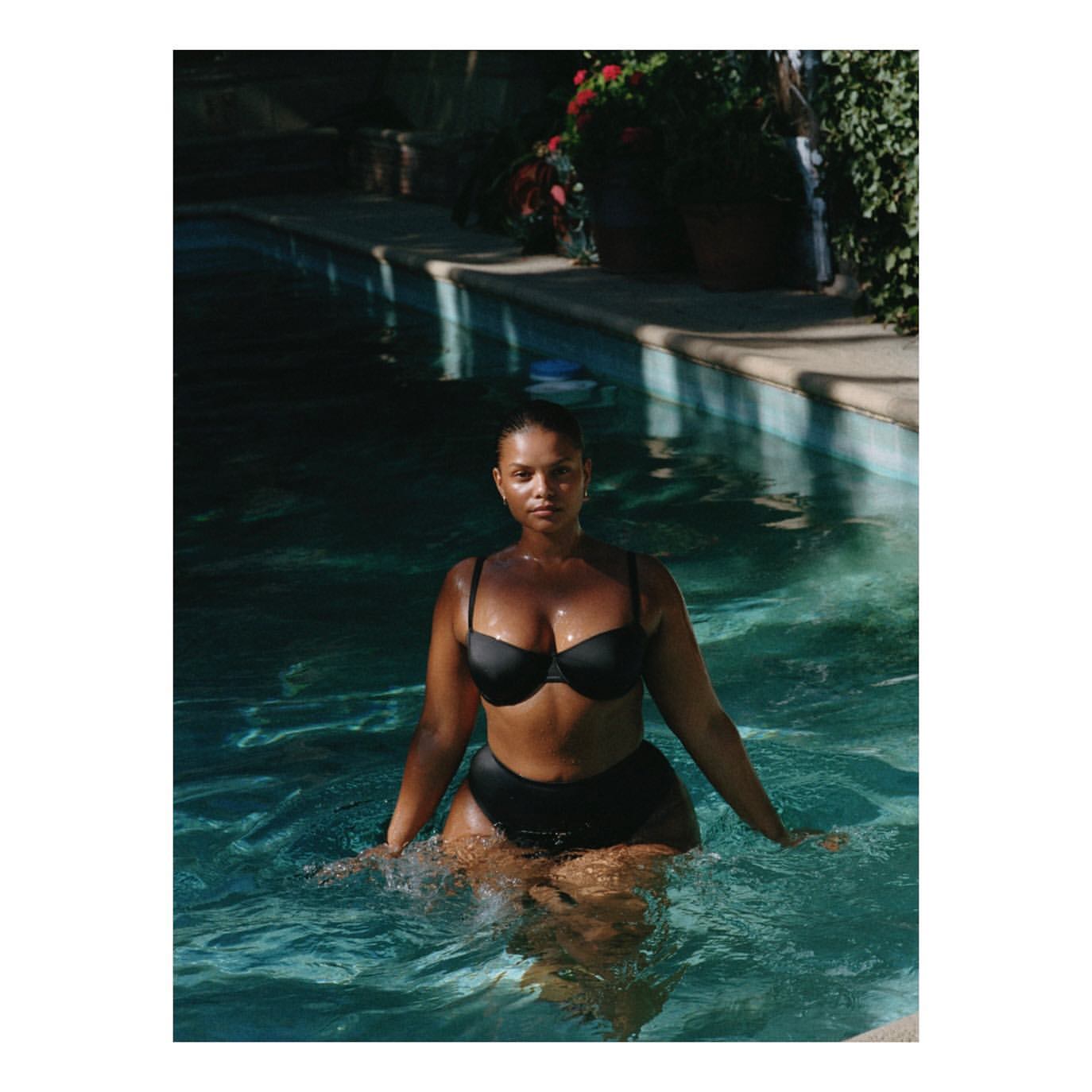 An adult woman is chest-deep in a pool, wearing a black bikini. She faces forward with a neutral expression on a sunny day.
