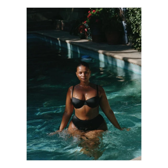 An adult woman is chest-deep in a pool, wearing a black bikini. She faces forward with a neutral expression on a sunny day.