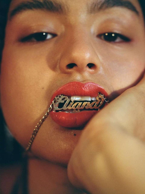 Close-up of a young woman with red lipstick, holding a gold necklace with the name 'Ciara' between her teeth, portrait orientation.