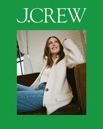 Julianne Moore seated on a brown sofa wearing a white jacket and blue jeans against an indoor brick wall.