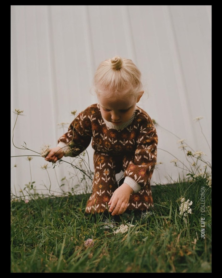 Toddler in brown floral print outfit plays with wildflowers in a grassy field against a white backdrop in a soft, dreamy outdoor setting.