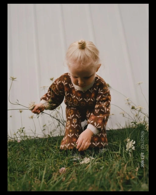 Toddler in brown floral print outfit plays with wildflowers in a grassy field against a white backdrop in a soft, dreamy outdoor setting.