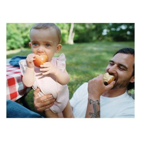 A baby eats an apple, held by an adult male also eating food, in an outdoor grassy setting with a checkered blanket.