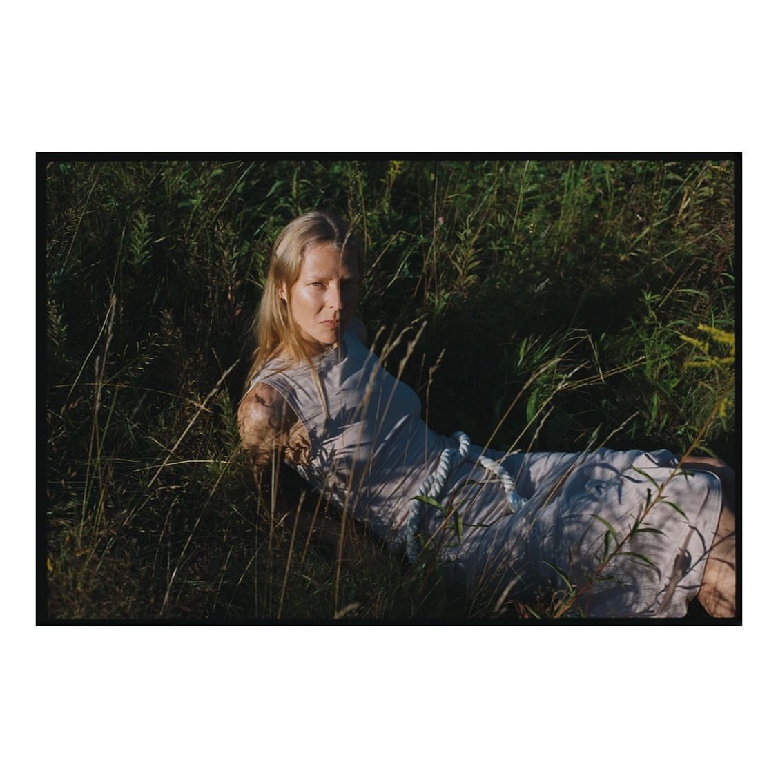 A woman with blonde hair lying in tall green grass wearing a light-colored dress with a white rope belt outdoors in a field setting.