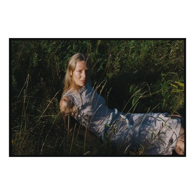 A woman with blonde hair lying in tall green grass wearing a light-colored dress with a white rope belt outdoors in a field setting.
