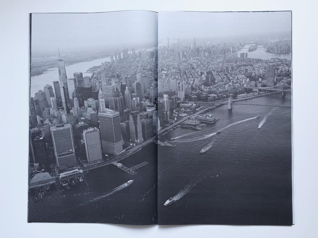 High-angle black and white photograph shows New York City skyline with tall buildings, waterways, and boat traffic.