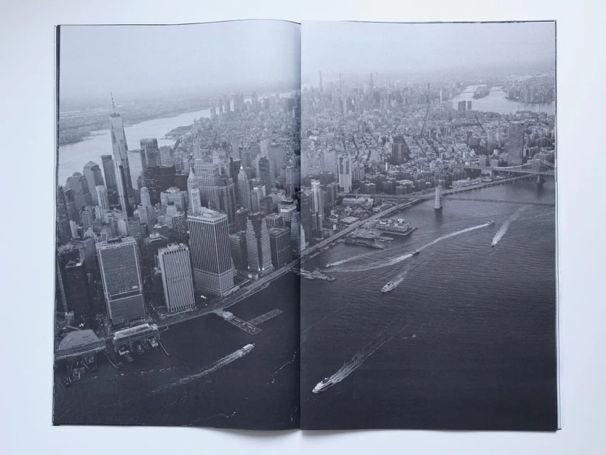 High-angle black and white photograph shows New York City skyline with tall buildings, waterways, and boat traffic.