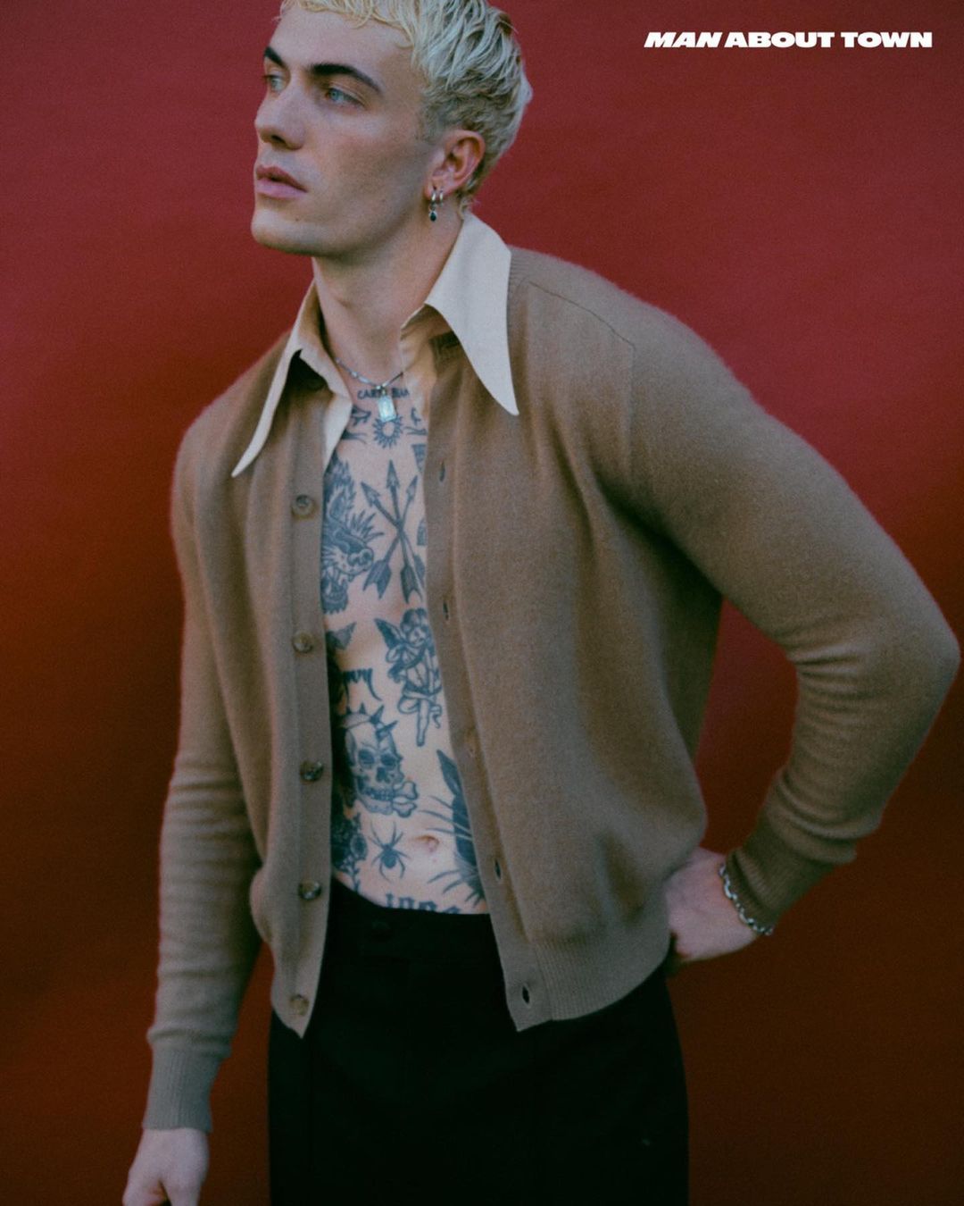 Portrait of a blond-haired man wearing a cardigan, posing against a red backdrop. He has tattoos and is wearing jewelry.