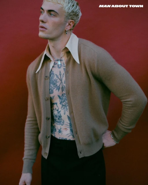 Portrait of a blond-haired man wearing a cardigan, posing against a red backdrop. He has tattoos and is wearing jewelry.