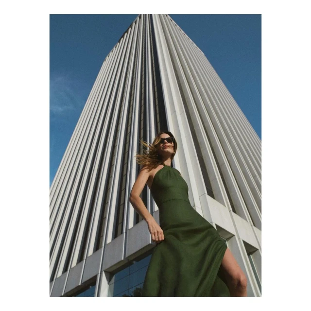 An adult woman in a green dress poses in front of a tall, modern, fluted building against a clear blue sky on a sunny day.