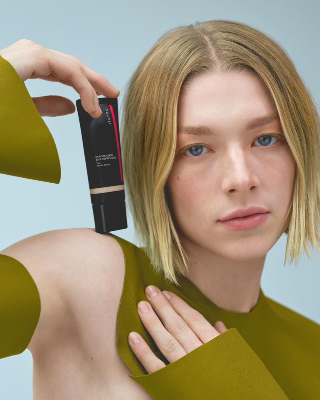 Woman holds a black tube of skin serum in a beauty product studio shot with golden shoulder-length hair, light skin, and blue eyes.