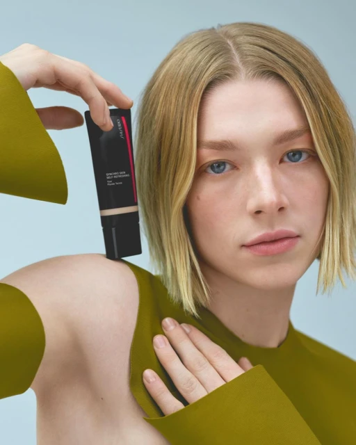 Woman holds a black tube of skin serum in a beauty product studio shot with golden shoulder-length hair, light skin, and blue eyes.