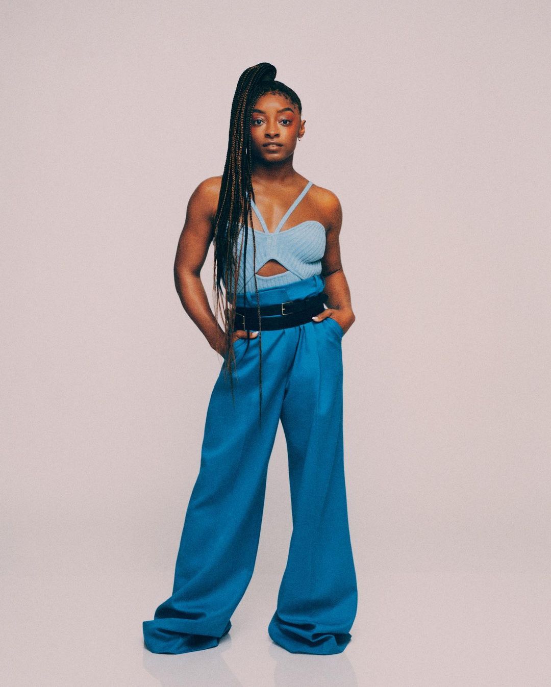 Full-length portrait of Simone Biles in a light blue knit top and blue palazzo pants, posed against a pale neutral studio backdrop.