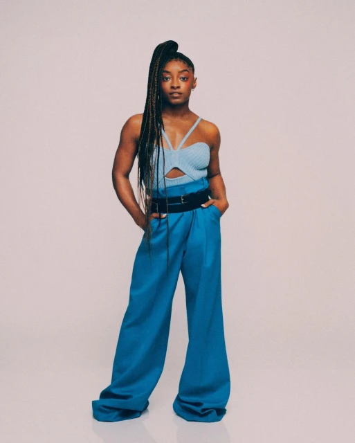 Full-length portrait of Simone Biles in a light blue knit top and blue palazzo pants, posed against a pale neutral studio backdrop.