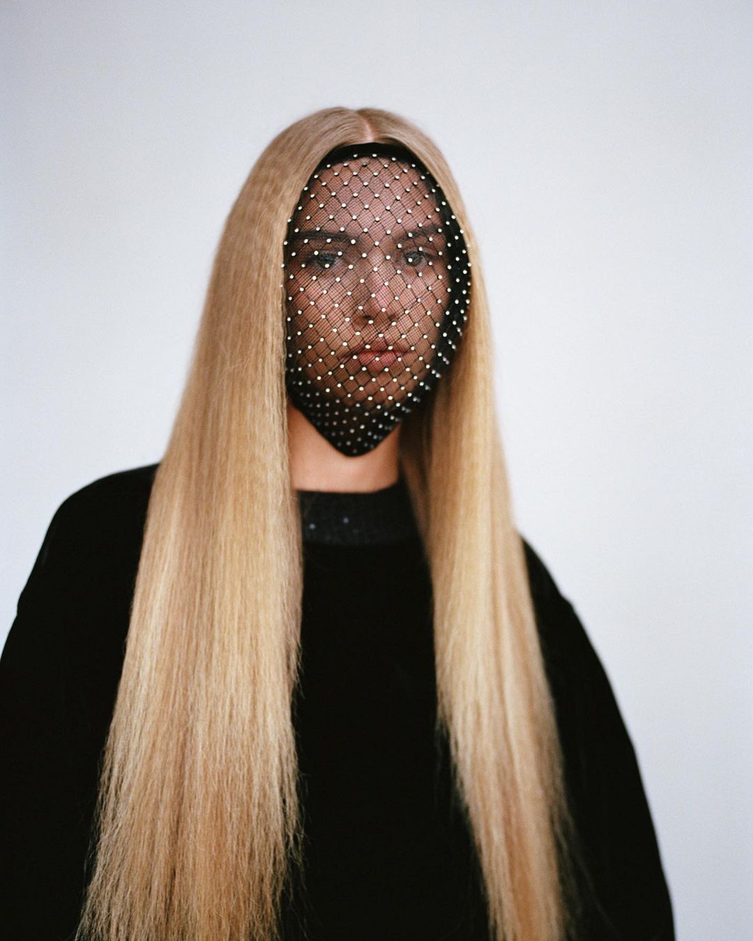 Woman with long blonde crimped hair and a studded face net poses against a white backdrop in a medium shot, fashion beauty portrait.