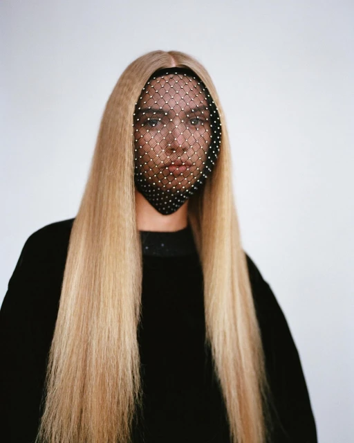Woman with long blonde crimped hair and a studded face net poses against a white backdrop in a medium shot, fashion beauty portrait.