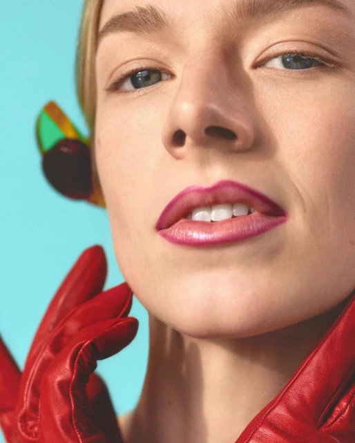 Close-up portrait of a fair-skinned woman with pink lipstick, wearing red leather gloves and colorful translucent earrings against a blue background.