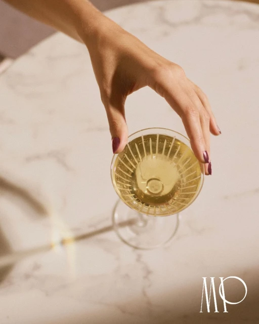 Overhead shot of a hand reaching for a glass of white wine on a round marble table with minimalist logo and soft, diffused lighting.