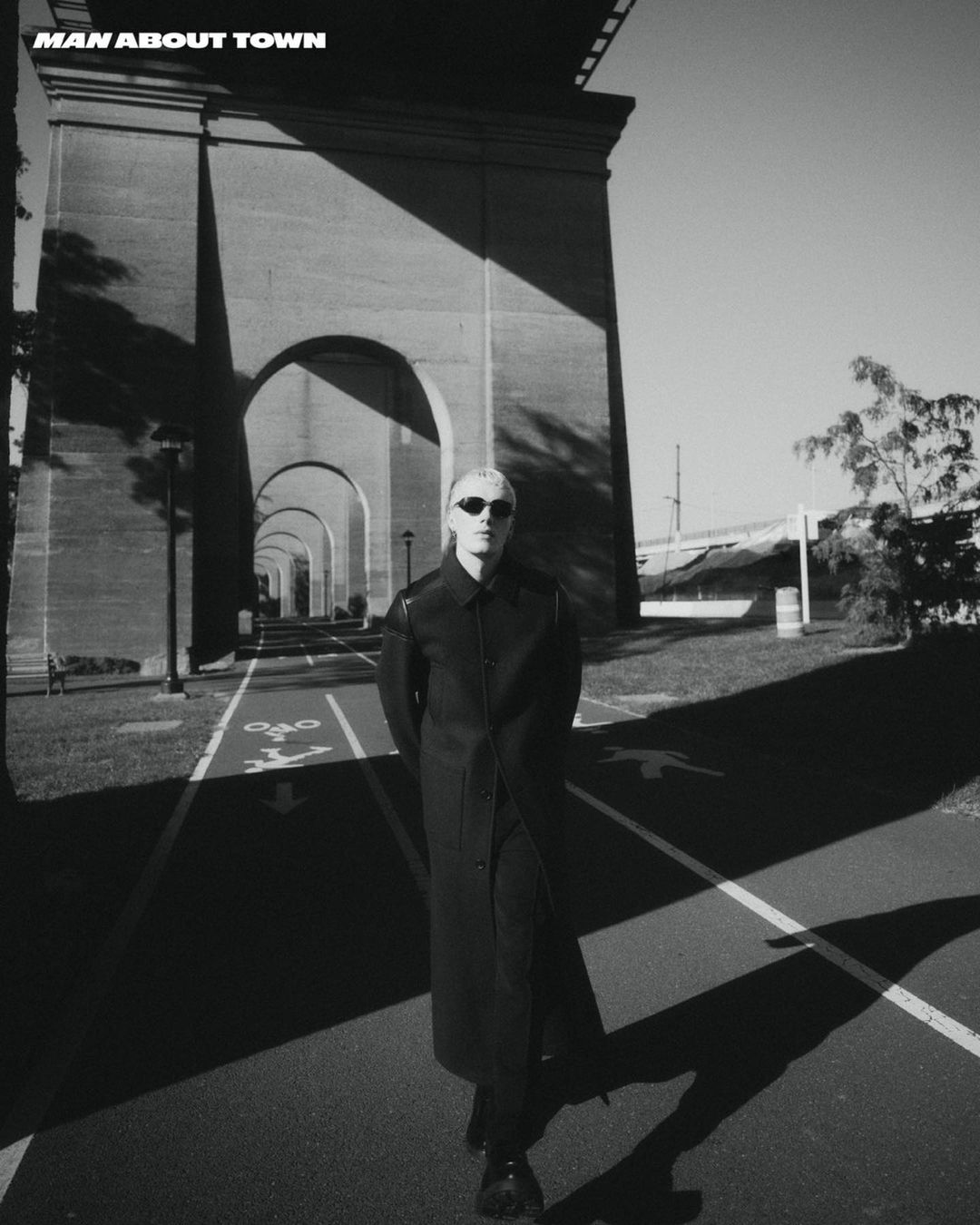 Man wearing sunglasses and a long dark coat walking beneath a large concrete bridge archway in New York