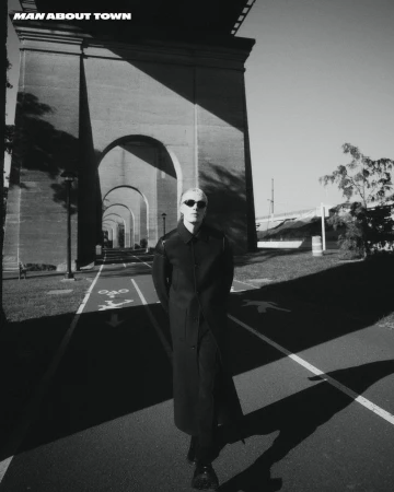 Man wearing sunglasses and a long dark coat walking beneath a large concrete bridge archway in New York