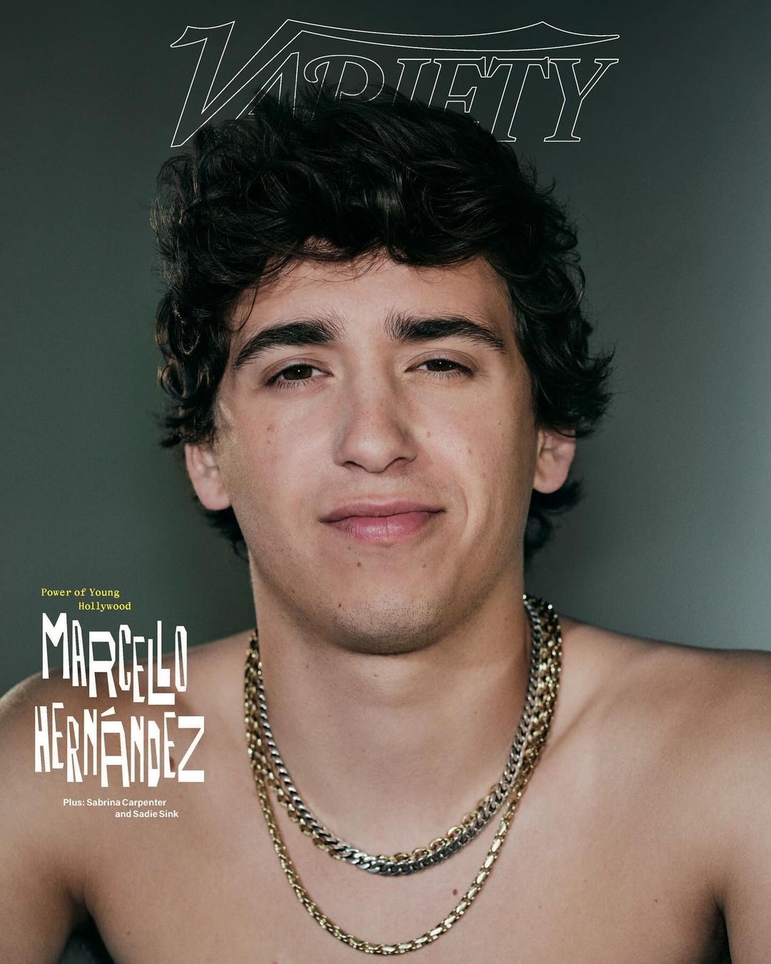 Portrait of young man with dark curly hair wearing layered silver and gold chains against a plain grey backdrop with magazine text overlays