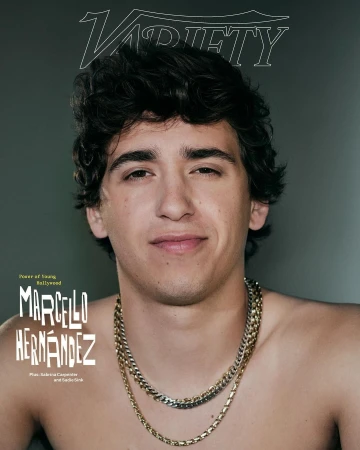 Portrait of young man with dark curly hair wearing layered silver and gold chains against a plain grey backdrop with magazine text overlays
