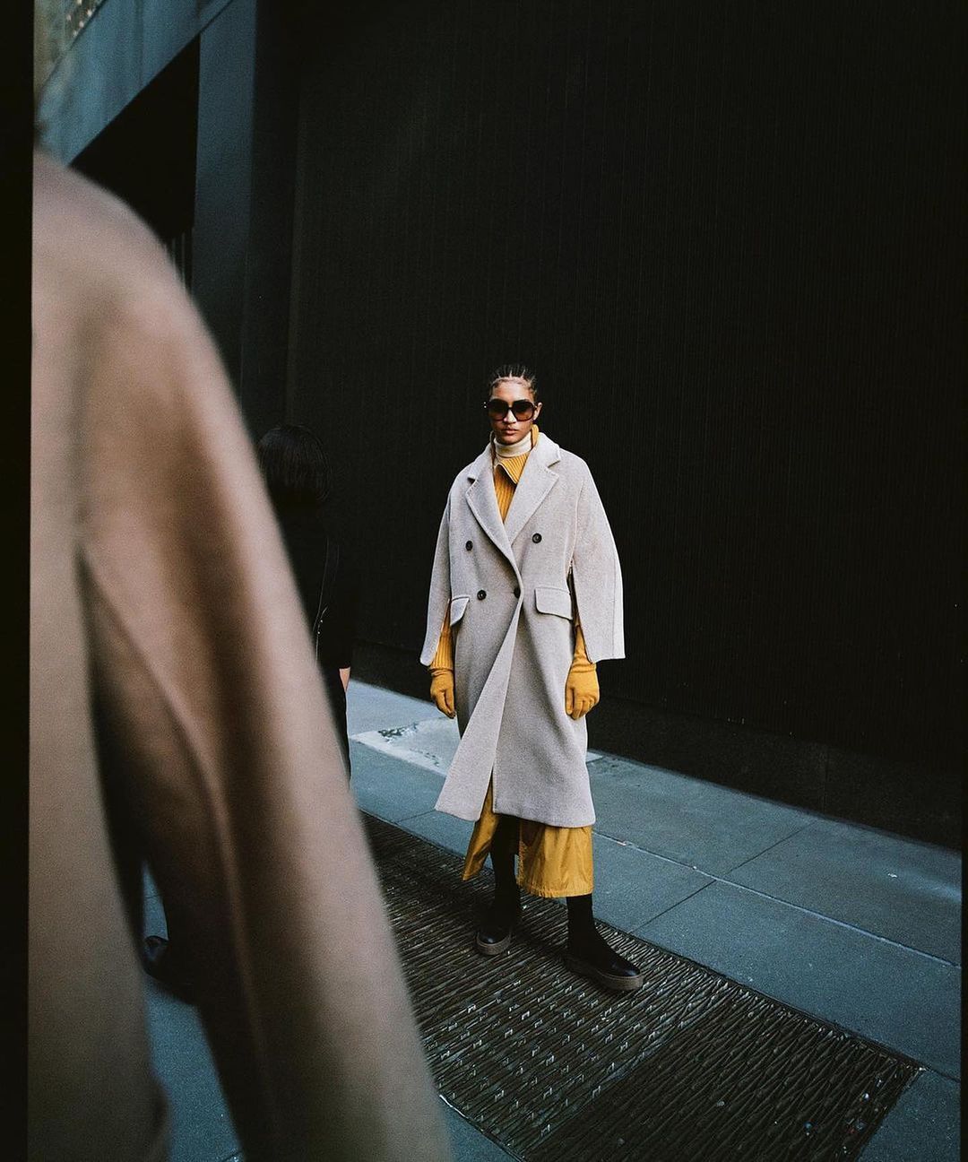 A woman in a tan coat and sunglasses walks confidently on an urban street in a fashion forward setting, exuding style and grace.