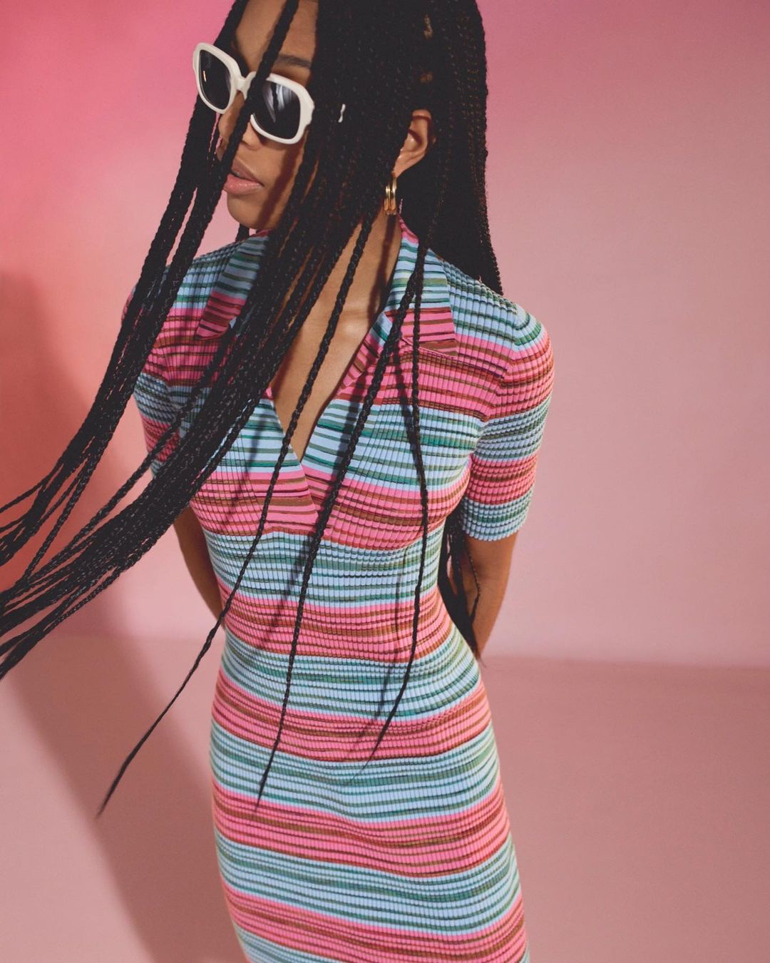 A young woman with long braids and sunglasses poses against a pink backdrop, wearing a multi-colored striped knit dress for a studio fashion portrait.