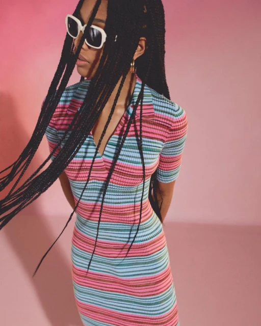 A young woman with long braids and sunglasses poses against a pink backdrop, wearing a multi-colored striped knit dress for a studio fashion portrait.
