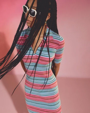A young woman with long braids and sunglasses poses against a pink backdrop, wearing a multi-colored striped knit dress for a studio fashion portrait.