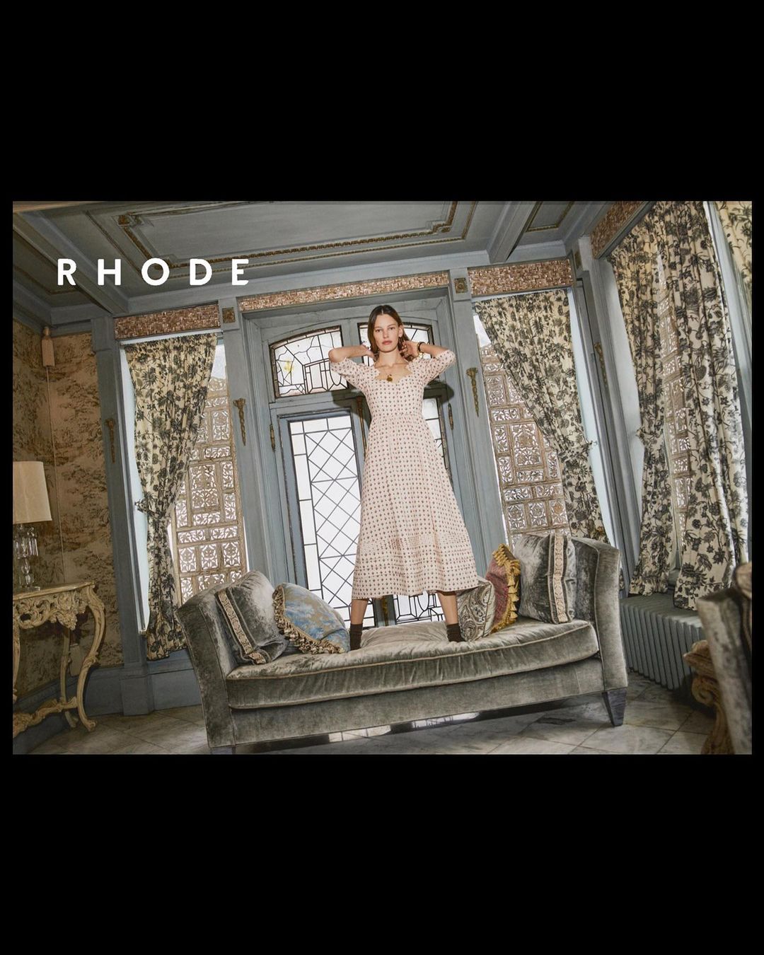 A woman in a long-sleeved dress standing on a tufted chaise lounge in front of a doorway with curtains and ornate walls.