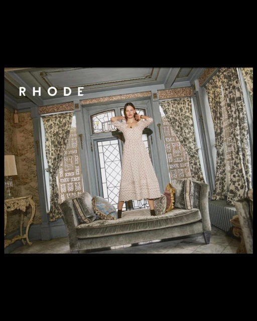 A woman in a long-sleeved dress standing on a tufted chaise lounge in front of a doorway with curtains and ornate walls.