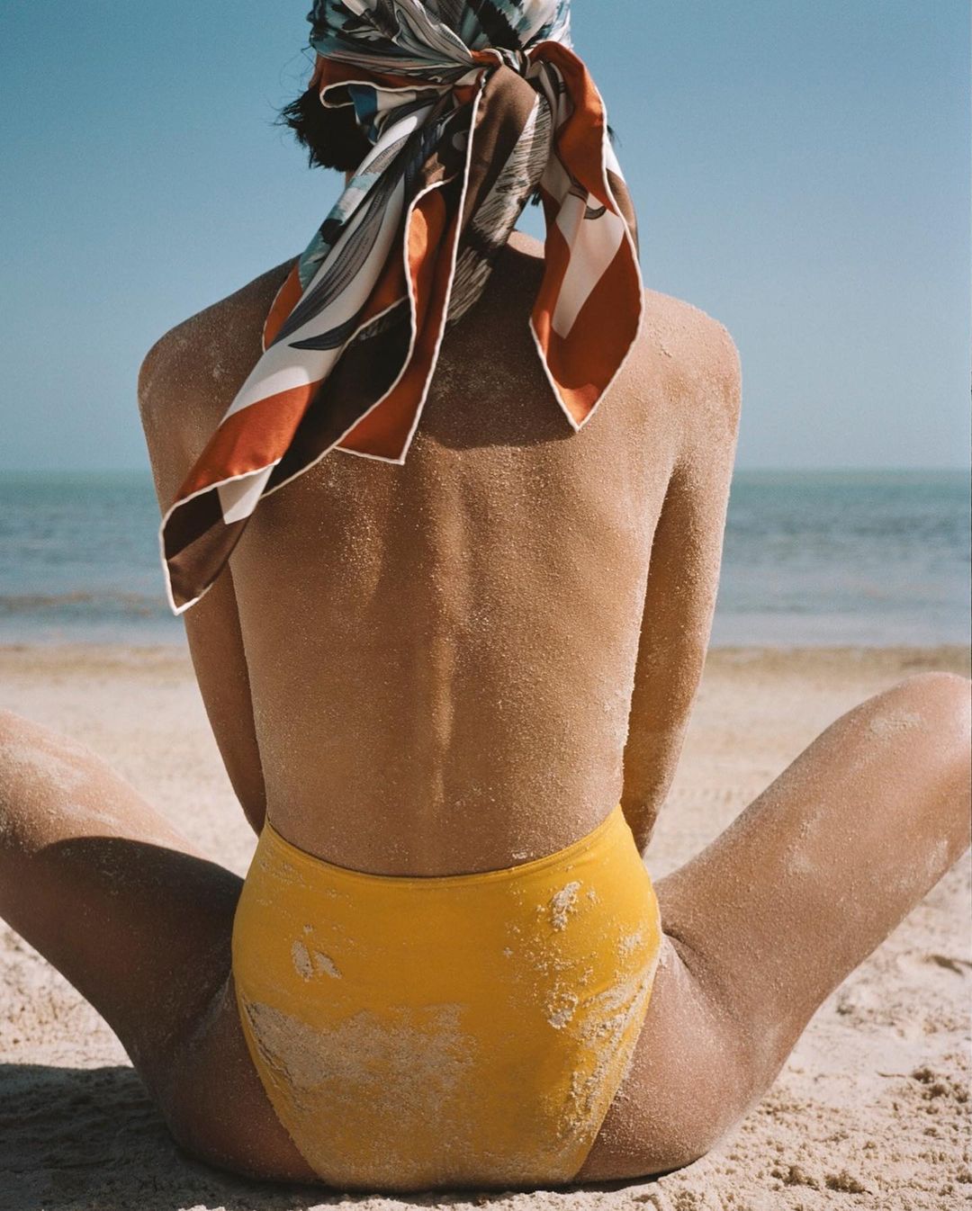 Back view of a woman in a yellow swimsuit sitting on a sandy beach with blue ocean and sky in the background wearing a patterned headscarf.