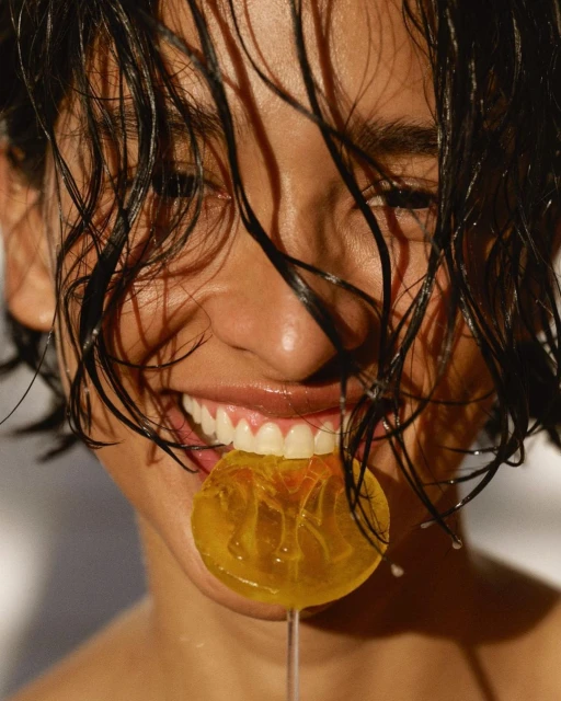 Close-up portrait of an adult female eating a yellow lollipop; she has wet, dark hair and is smiling with her teeth showing against a white background.