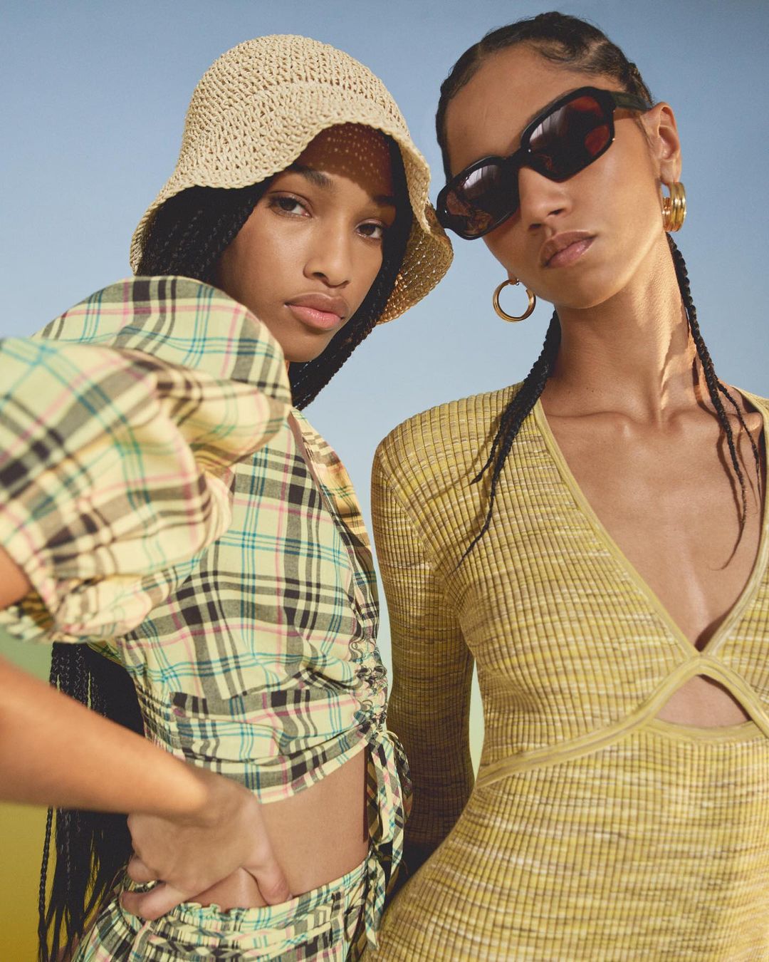 Two adult females wearing fashionable yellow clothing and black sunglasses in a medium shot, posing in an outdoor setting with a blue sky.