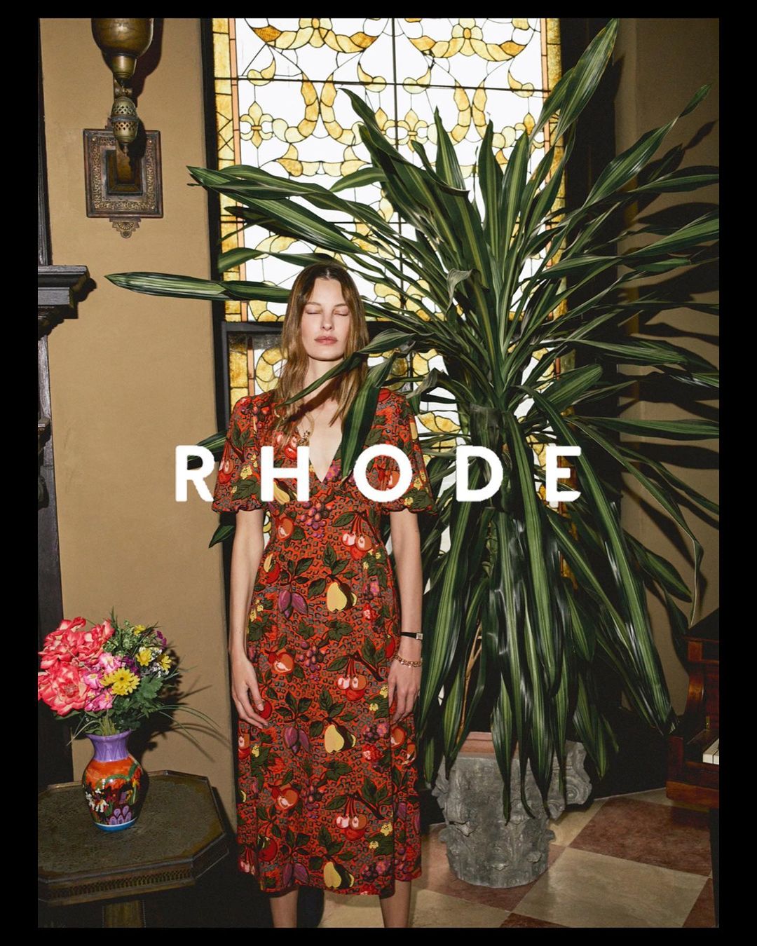 A woman poses in a reddish-brown floral dress next to a potted plant and a window with geometric stained-glass.