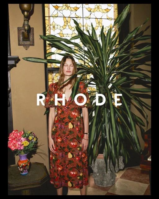 A woman poses in a reddish-brown floral dress next to a potted plant and a window with geometric stained-glass.