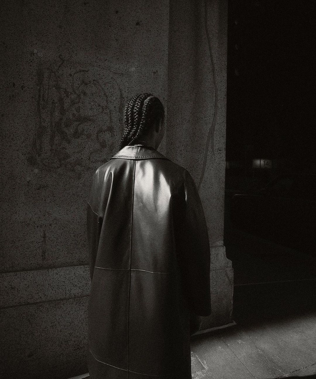 Back view of a person in a black leather coat standing next to a marble wall in a dimly lit indoor environment. Fashion editorial.