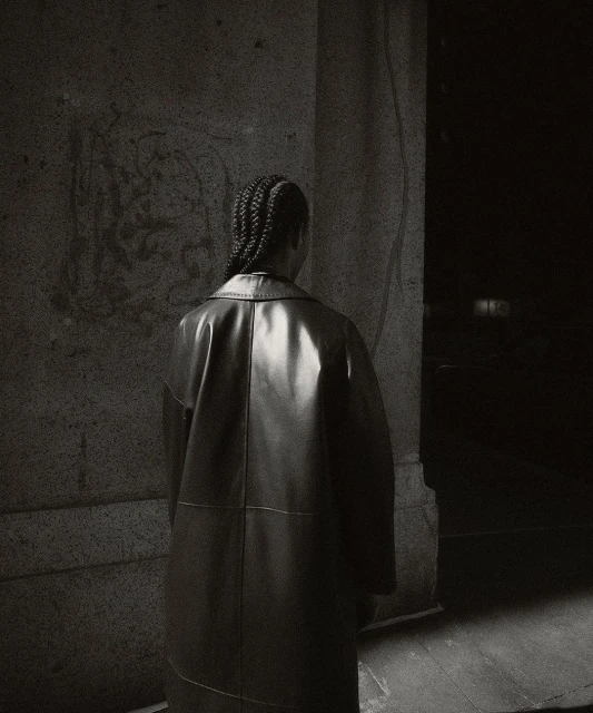 Back view of a person in a black leather coat standing next to a marble wall in a dimly lit indoor environment. Fashion editorial.