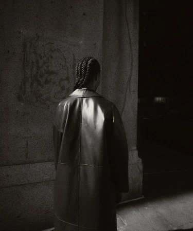 Back view of a person in a black leather coat standing next to a marble wall in a dimly lit indoor environment. Fashion editorial.