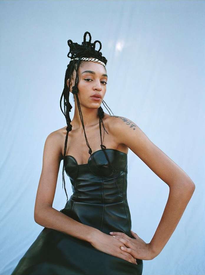 Portrait of an adult woman with braided hair, wearing a black leather dress and a silver headpiece, posing against a light blue background.