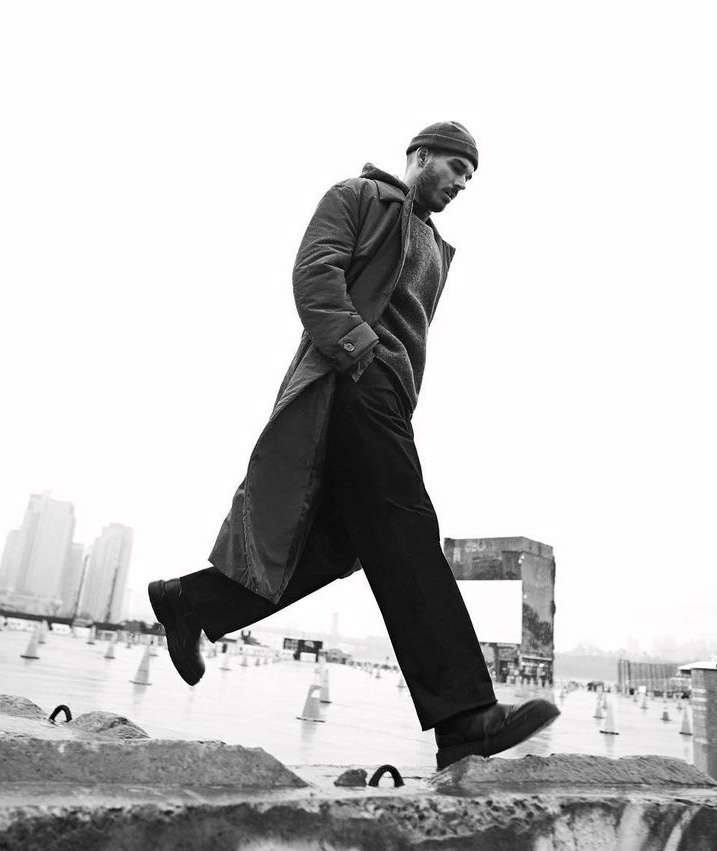 Monochrome portrait of a man in a coat and beanie walking on a ledge against an urban background. Full body shot, low angle.