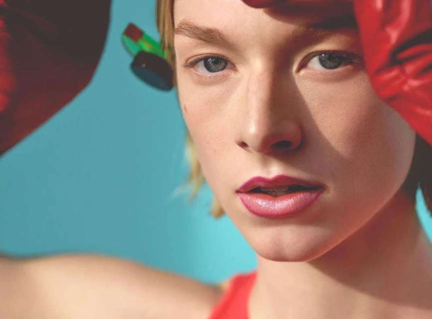 Close-up portrait of a young adult female with fair skin, striking blue eyes, and red boxing gloves against a vibrant turquoise backdrop.