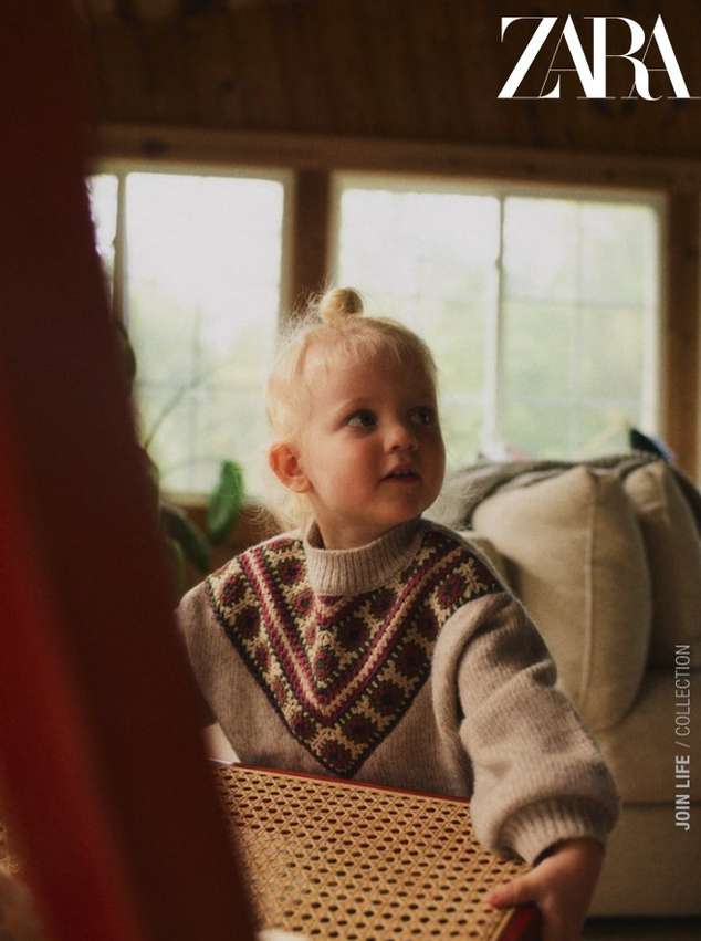 Young girl with blond hair in a bun sits at a wicker chair in front of a window, wearing a patterned sweater. Zara logo visible.