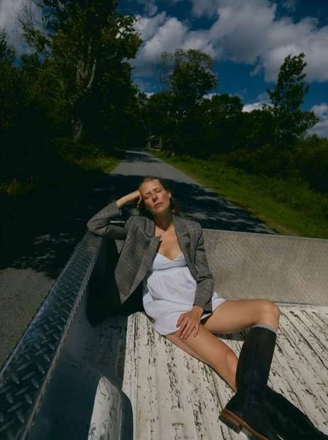 Woman in blazer and dress sits on a truck bed on a road with trees and sky in the background, creating a relaxed, outdoor fashion scene.